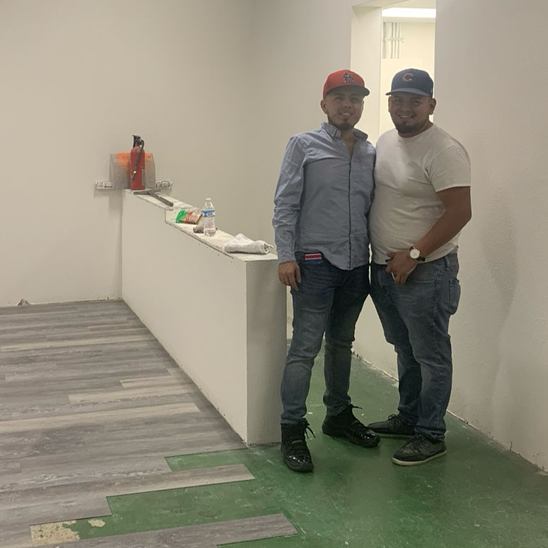 Two men stand together in a partially finished room with exposed flooring; supplies and a fire extinguisher are on a counter behind them.